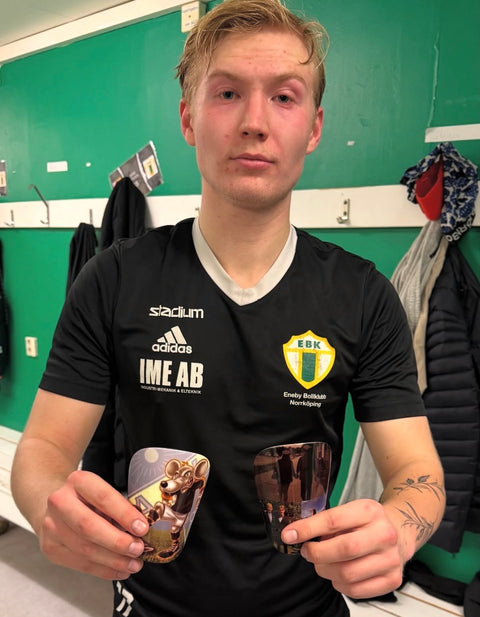 Fotbollsspelare i svart klubbtröja håller upp personliga fotbollsbenskydd med egna bilder